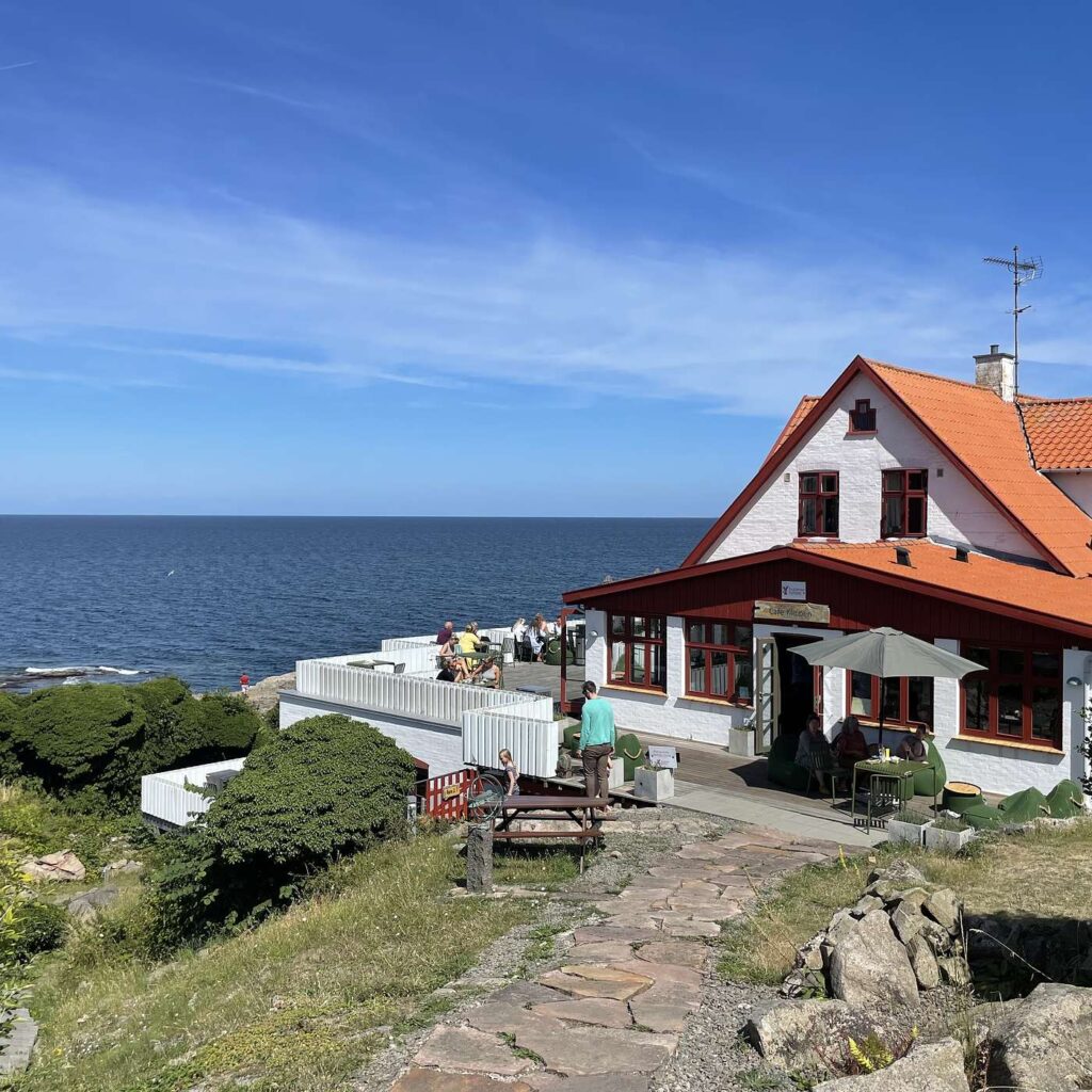 Et hus med rødt tag og en terrasse ved havet under en blå himmel.