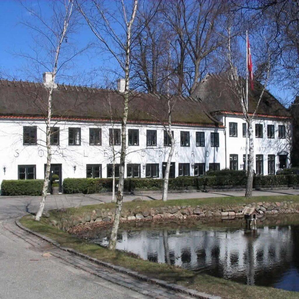 En hvid bygning ved en s&oslash; med spejling i vandet og tr&aelig;er langs bredden.