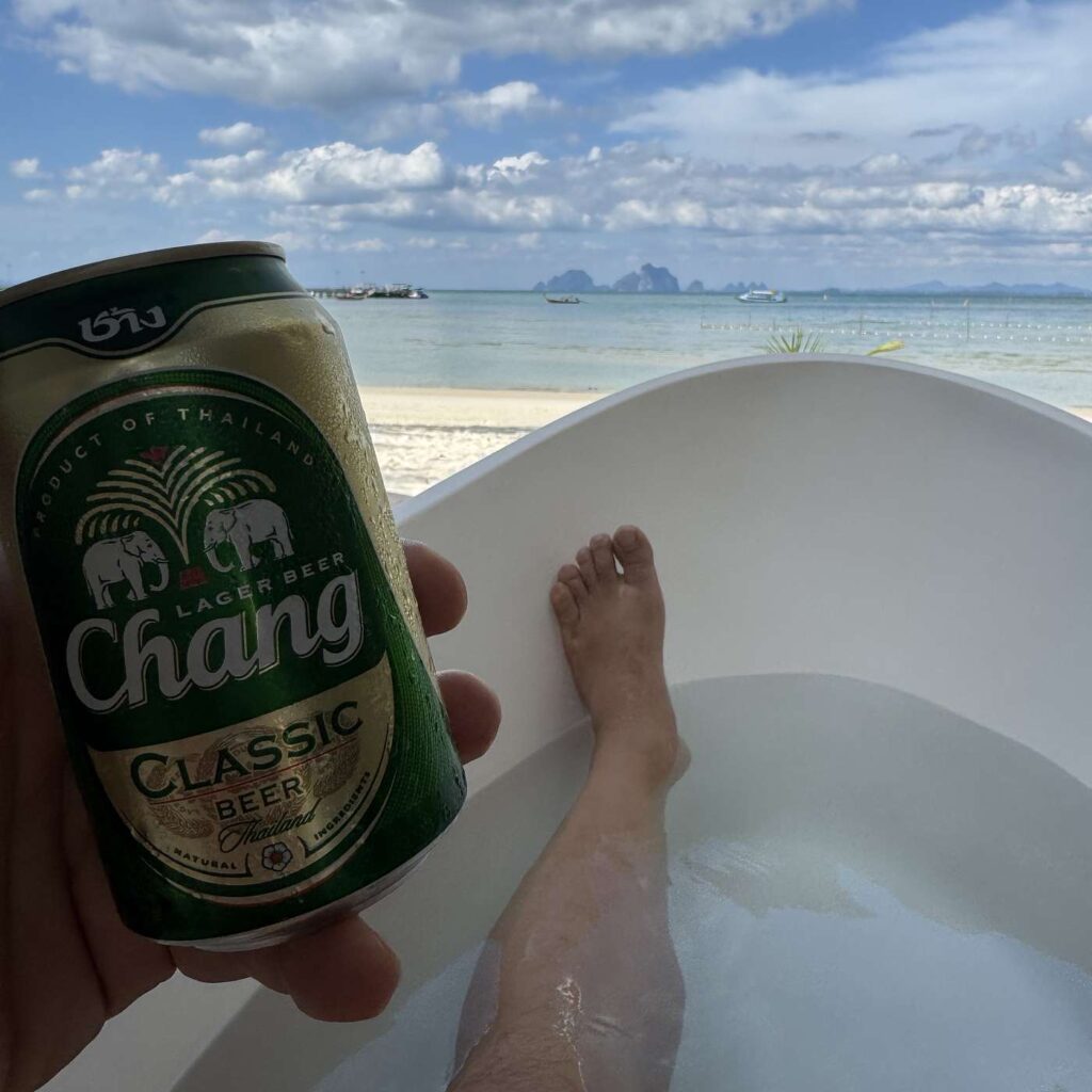 En hånd holder en Chang-øldåse foran et badekar med udsigt over havet.