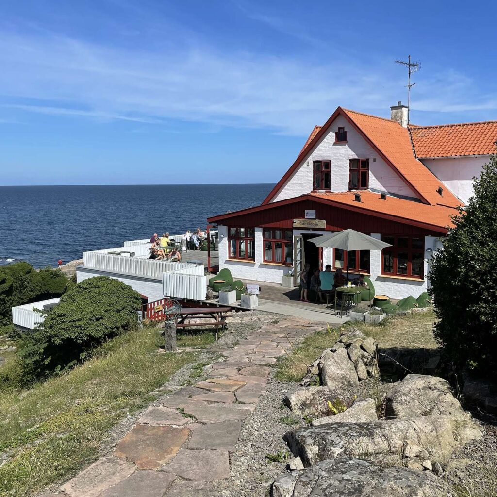 Et hus med r&oslash;dt tag og en terrasse ved havet under en bl&aring; himmel.